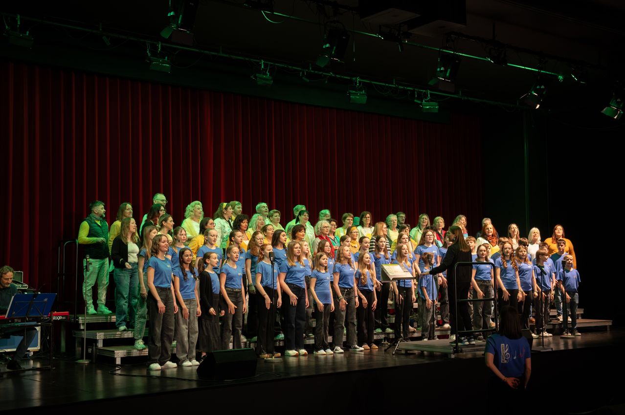Ein Chor, in passenden blauen T-Shirts gekleidet, tritt auf einer Bühne mit roten Vorhängen und grünen Bühnenlichtern auf. Das Publikum sitzt auf der Bühne, und eine Frau steht auf einem Wagen mit Notenblättern.