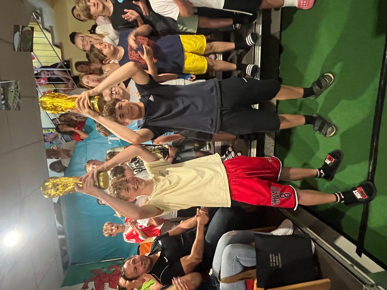 Eine Gruppe junger Jungen feiert das Hochhalten eines Pokals in einem Raum. Sie lächeln und scheinen in einem Wettbewerb zu sein.