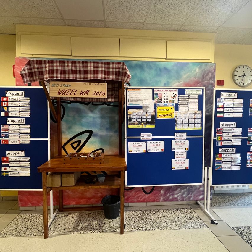Ein Holzstand mit rotem und weißem Vordach zeigt Informationen über ein Fußballereignis. Der Stand enthält eine Holzbox mit Gegenständen darin. Mehrere Tafeln mit Gruppennamen und Flaggen sind an der Wand montiert.