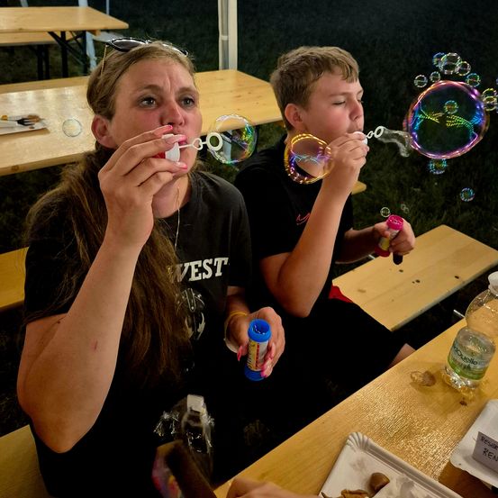 Eine Frau und ein Junge blasen mit Blasenstäben Seifenblasen und sitzen an einem Picknicktisch mit Flaschen und Tellern in der Nähe.