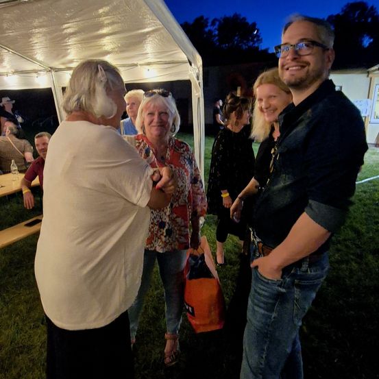 Eine Gruppe von Menschen steht bei Nacht unter einem Zelt. Eine Frau in Weiß gibt einer Frau in einem Blumenhemd die Hand. Ein Mann mit Brille und Gürtel lächelt.