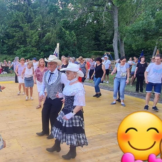 Ein Paar in Cowboy-Kleidung tanzt auf einem Holzboden, umgeben von einer Menschenmenge, Bäumen und einer Fahne.