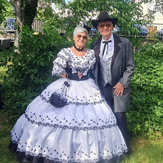 Ein älteres Paar steht vor einem grünen Busch. Die Frau trägt ein weißes Kleid mit schwarzem Besatz und einem Hut. Der Mann trägt einen Anzug und einen Hut.