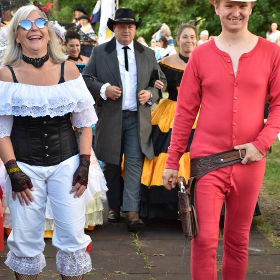 Eine Gruppe von Menschen in Kostümen geht auf einem Bürgersteig. Eine Frau in einem Korsett lächelt, während ein Mann in einem roten Outfit neben ihr steht. Hinter ihnen bewegen sich Menschen in verschiedenen Kostümen.
