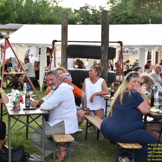 Mehrere Erwachsene sitzen an Tischen auf einem Open-Air-Festival. Ein Mann hat ein Bier, und eine Frau lächelt. Der Bereich ist mit Tischen und Zelten gefüllt.