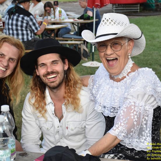 Eine Gruppe von Menschen bei einer Veranstaltung im Freien, lächelnd. Ein Mann in einem Cowboyhut und eine Frau in einem weißen Kleid und Hut lachen zusammen. Flaschen und ein Tisch sind im Vordergrund.