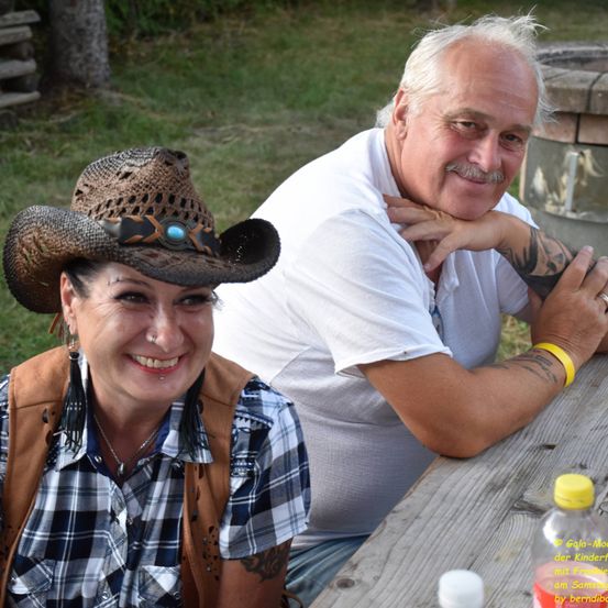 Eine lächelnde Frau mit einem Cowboyhut und ein Mann mit Tattoos sitzen gemeinsam an einem Picknicktisch. Der Mann hat ein gelbes Armband und einen Ring. Auf dem Tisch stehen eine Flasche und ein Becher.