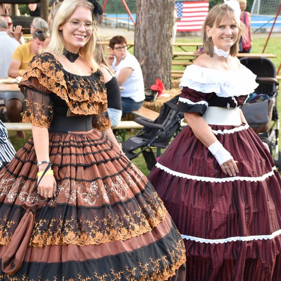 Zwei Frauen in Vintage-Kleidern stehen bei einem Treffen draußen. Die Frau auf der linken Seite trägt ein braunes Kleid mit Spitze. Die Frau auf der rechten Seite trägt ein burgunderrotes Kleid. Hinter ihnen sitzen Leute an Picknicktischen und einem Baum.