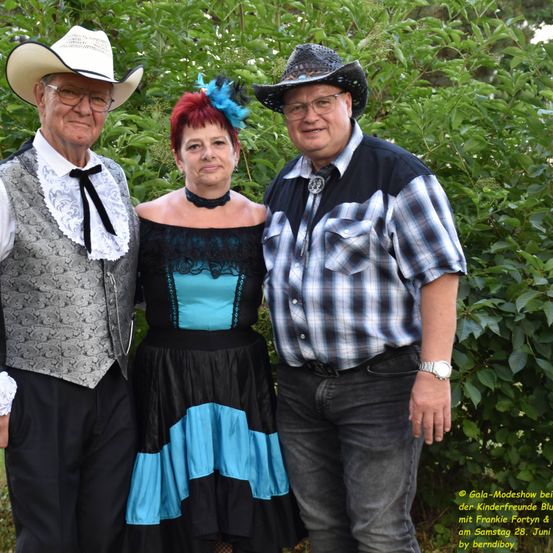Drei Personen in Western-Kleidung posieren für ein Foto vor grünem Laub. Der Mann links trägt einen Cowboyhut, eine Weste und eine Fliege. Die Frau in der Mitte hat eine blaue Feder im Haar. Der Mann rechts trägt einen Cowboyhut, ein kariertes Hemd und eine Uhr.
