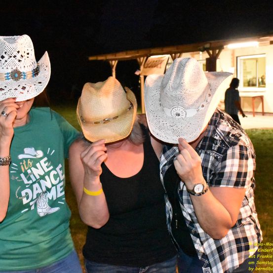 Drei Menschen mit Cowboy-Huten, zwei Frauen und ein Mann, posieren für ein Foto auf einer Rasenfläche bei Nacht.