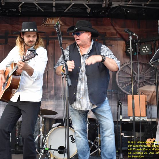 Zwei Männer treten auf einer Bühne auf. Einer spielt Gitarre, der andere singt. Dahinter steht ein Schlagzeug und eine Kulisse mit hölzernem Design. Das Bild ist von Gala-Modeshow gekennzeichnet.