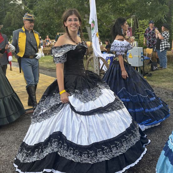 Eine Frau in einem schwarz-weißen Kleid steht im Freien. Sie lächelt in die Kamera. Hinter ihr stehen eine weitere Frau und ein Mann in Uniform. Im Hintergrund befinden sich Bäume und eine weiße Flagge.
