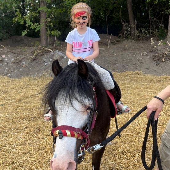 Ein junges Mädchen mit Gesichtsbemalung reitet ein kleines Pferd auf einem Feld. Eine Person hält die Zügel. Bäume und Gras umgeben das Gebiet.