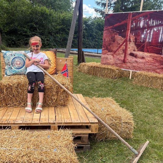 Ein junges Mädchen sitzt auf Heuballen und hält eine Harke. Sie hat Gesichtsbemalung und posiert für ein Foto. Dahinter befindet sich ein großes Bild eines Bauernhofs.