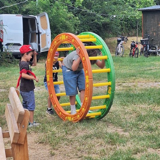 Drei Jungen spielen auf einem bunten Ringspielzeug auf einer Wiese. Ein Junge klettert darauf, während die anderen beiden zusehen. Ein weißer Lieferwagen und Fahrräder sind in der Nähe geparkt.