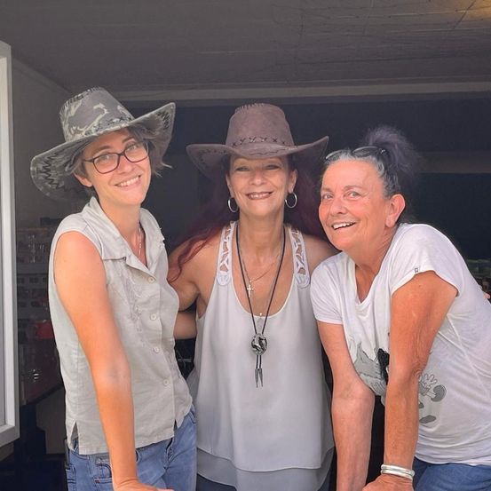 Drei Frauen lächeln in einem Raum, zwei tragen Cowboyhüte und eine mit Brille. Die Frau in der Mitte trägt eine Halskette mit einem Kreuz. Sie scheinen für ein Foto zu posieren.