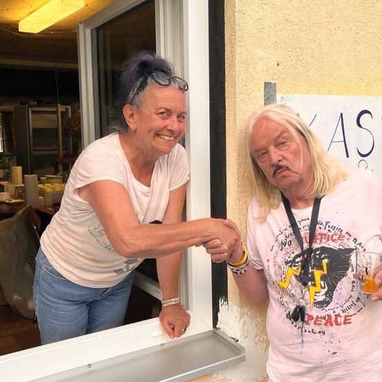 Zwei Erwachsene stehen lächelnd vor einem Fenster und schütteln sich die Hände. Eine Frau hat eine Brille auf dem Kopf und ein Mann hält ein Getränk. Hinter ihnen befindet sich ein Banner mit Text.