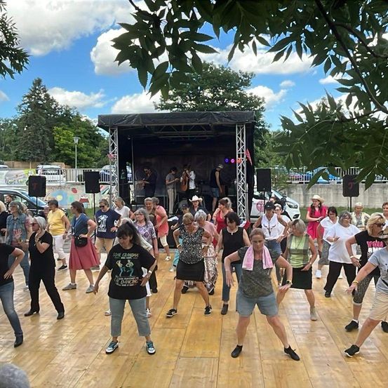 Eine Gruppe älterer Frauen tanzt auf einer Holzbühne in einer Freiluftumgebung. Im Hintergrund ist eine Bühne mit einer Band zu sehen. Die Frauen tragen lässige Sommerkleidung.