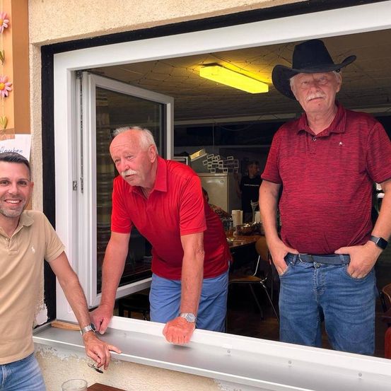 Drei Männer posieren vor einem Fenster. Einer lehnt sich auf die Fensterbank, ein anderer schaut nach draußen, und der dritte steht mit den Händen in den Hosentaschen. Ein Kühlschrank und ein Tisch mit Stühlen sind durch das Fenster sichtbar.