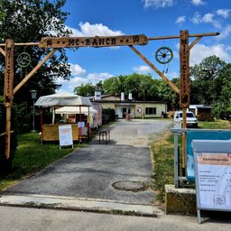 Der Eingang zum MC Ranch ist ein hölzerner Torbogen mit einem Holzschild und einem Traumfänger, der daran hängt. Es gibt Zelte, einen Tisch und ein Hinweisschild auf der Seite. Ein Gebäude mit einem Schornstein und ein geparkter Van befinden sich dahinter.