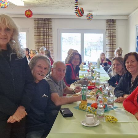 Eine Gruppe von Menschen sitzt an einem langen Tisch mit grüner Tischdecke und lächelt. Flaschen, Tassen und Teller stehen auf dem Tisch. Eine Frau steht hinter ihnen, und ein Mann sitzt davor. Dahinter ist ein Raum mit Glastür und vorhängenbehangenen Fenstern.