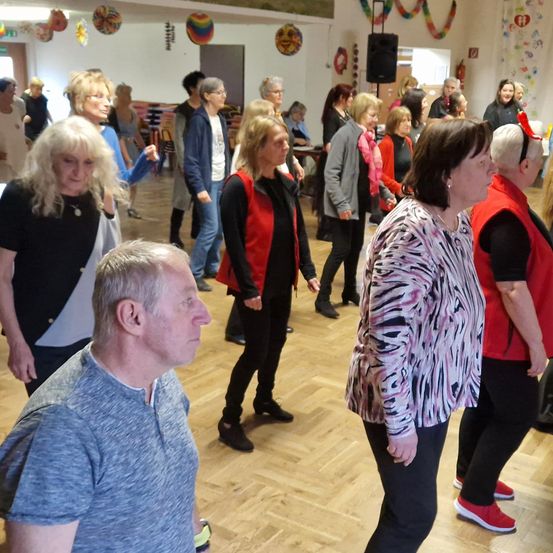 Eine Gruppe von Menschen tanzt in einem Raum mit Holzböden. Einige tragen Brillen und Turnschuhe. Eine Frau in einer roten Weste schaut den vor ihr stehenden Mann an. Der Raum hat einen Lautsprecher und bunte Dekorationen hängen an der Wand.