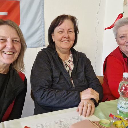 Drei Frauen sitzen an einem Tisch und lächeln für ein Foto. Im Hintergrund hängen eine Schweizer Flagge und eine rote Chilischote.
