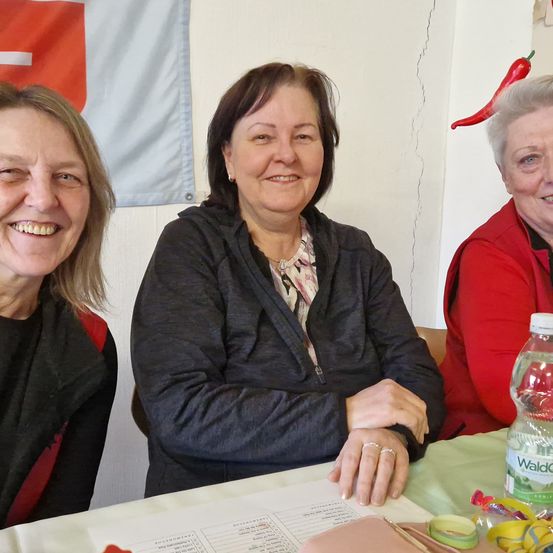 Drei Frauen sitzen an einem Tisch und lächeln für ein Foto. Im Hintergrund hängen eine Schweizer Flagge und eine rote Chilischote.