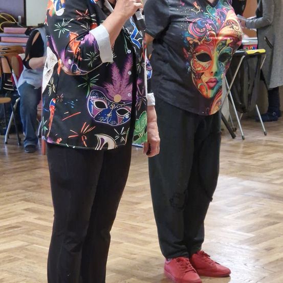 Zwei Frauen, eine mit einem Mikrofon, stehen zusammen auf einem Holzboden. Sie tragen bunte Oberteile und schwarze Hosen. Dahinter steht eine Person in einem grauen Mantel in der Nähe eines Tisches.