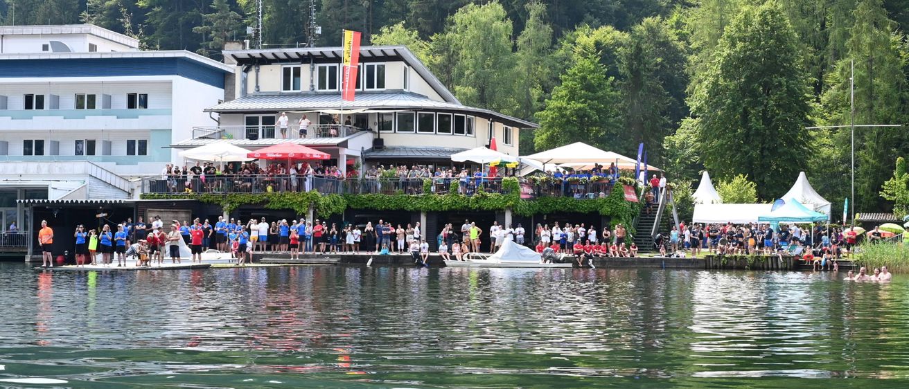 Eine Menschenmenge versammelt sich am See in der Nähe eines Hauses mit einer roten Fahne. Leute stehen auf einer Pier, einige in blauen Shirts, mit Booten im Wasser.