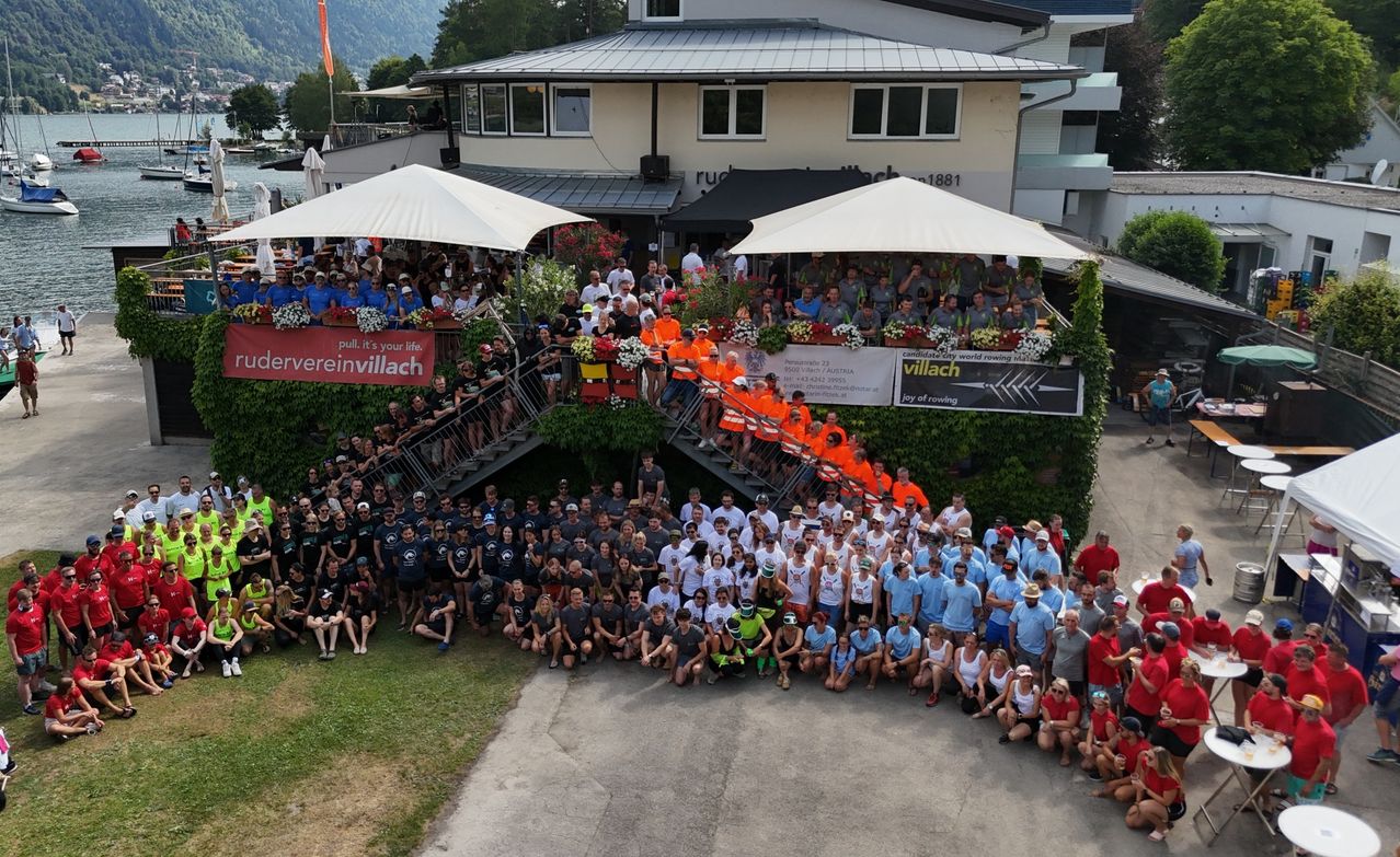 Gruppenfoto von Menschen in blauen und orangefarbenen Shirts bei einer Ruder-Weltmeisterschaft in Villach. Ein Gebäude mit Balkon und Treppen steht hinter ihnen.