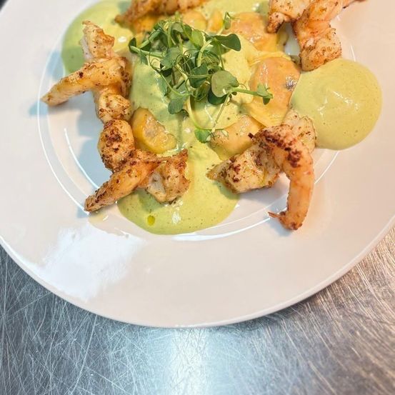 Ein weißer Teller mit gebratenen Garnelen, grüner Sauce und Kräutergarnitur, serviert auf einer metallischen Oberfläche.