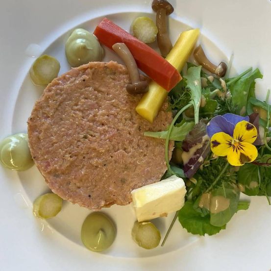 Ein Teller zeigt eine Scheibe Fleisch, ein Stück Käse, einen Salat mit Blättern und einer gelben Blume sowie einige grüne und braune Pilze.