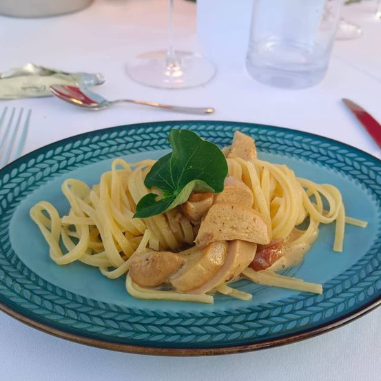 Ein Teller mit Pasta und Hähnchen, garniert mit einem grünen Blatt, steht auf einem Tisch mit Besteck und einem Weinglas.