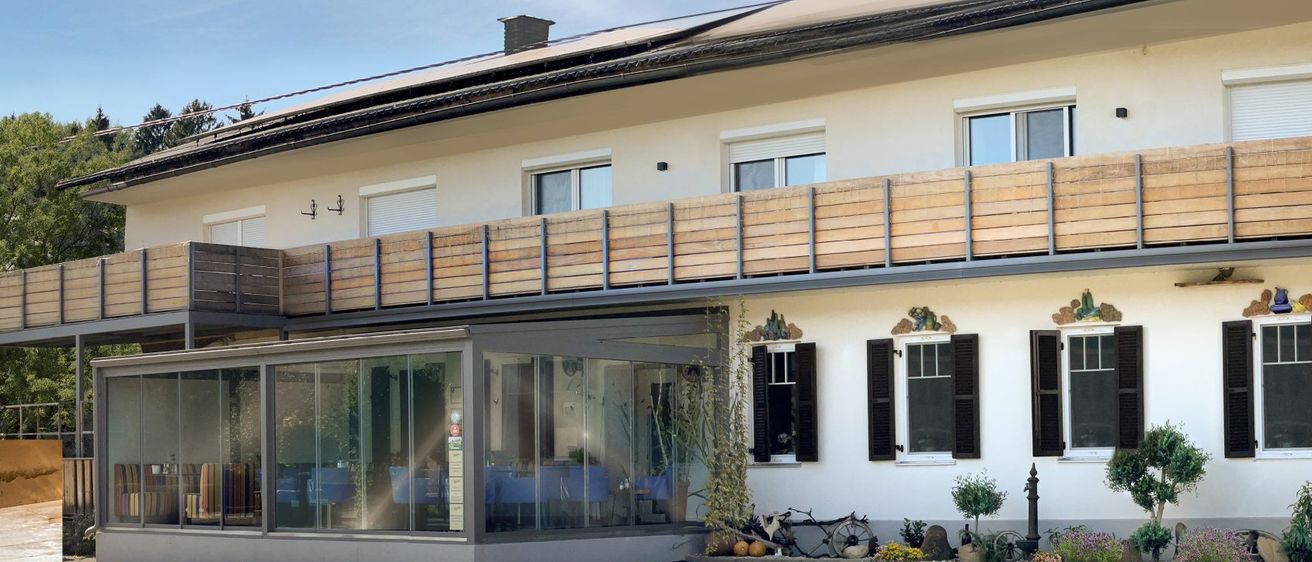 Das Bild zeigt ein modernes weißes Gebäude mit Solarmodulen auf dem Dach, Glasfenstern und einem Balkon mit Holzgeländern. Es gibt einen Swimmingpool, Liegestühle und Bäume im Hintergrund.