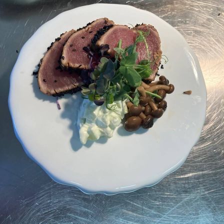 Ein weißer Teller zeigt ein Gericht aus gebratenem Thunfisch, garniert mit Pilzen und Grünzeug, dazu eine cremige weiße Sauce.