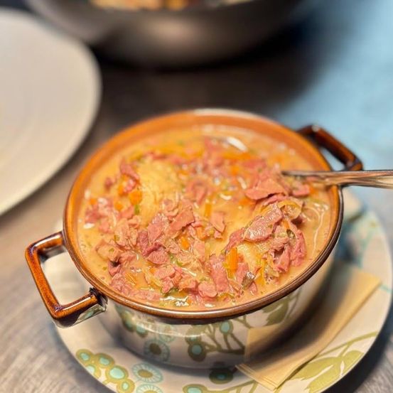 Eine Schüssel mit herzhafter Suppe mit Schinken und Gemüse auf einem Blumenteller, begleitet von einem Löffel und einem unscharfen Hintergrund.