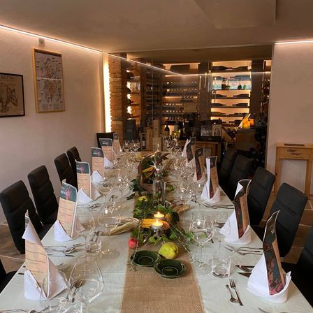 Ein langer Tisch für eine Mahlzeit mit Weinläsern, Tellern und Servietten. Dahinter Regale mit Flaschen, eine Ziegelwand und eine Karte an der Wand.
