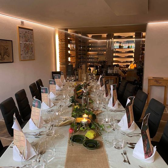 Ein langer Tisch für eine Mahlzeit mit Weinläsern, Tellern und Servietten. Dahinter Regale mit Flaschen, eine Ziegelwand und eine Karte an der Wand.