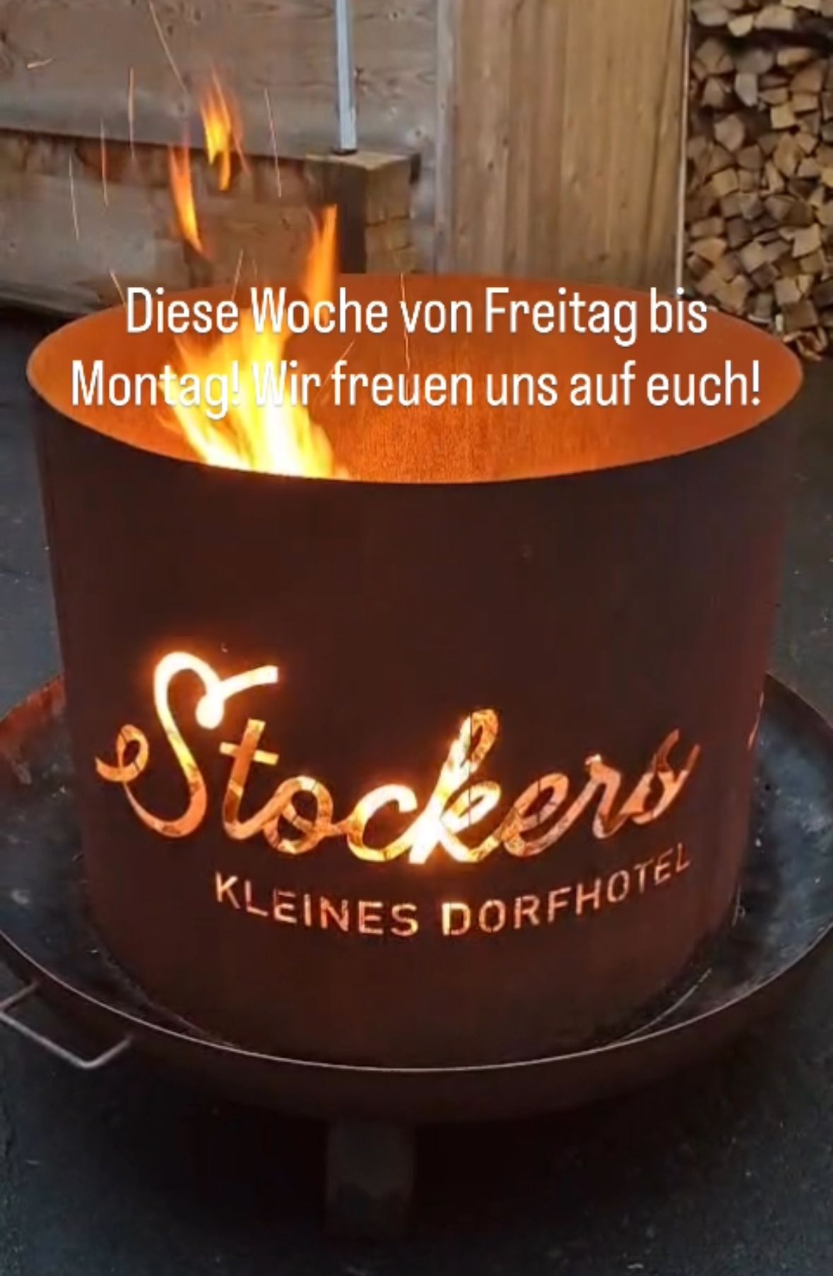 A fire pit with a burning fire inside. The words 'Stockers Kleines Dorfhotel' are written on the side. The fire pit is lit up with a bright light. There are words written on top of the fire pit.