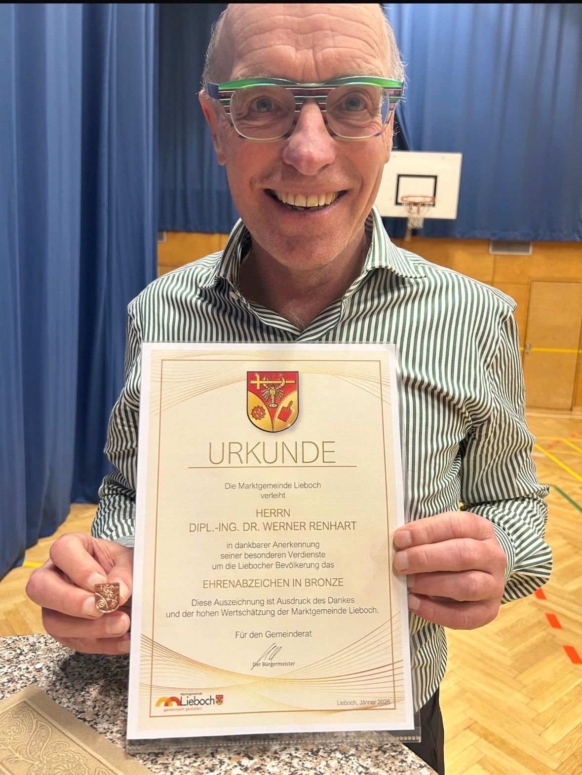 A man holding a certificate with a bronze medallion, smiling in a gymnasium. Behind him, a basketball hoop and a blue curtain.