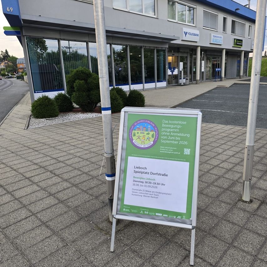 A signboard with green and white text is placed in front of a building. The sign reads 'Lieboch Spielplatz Dorfstraße' with details about the location and date.