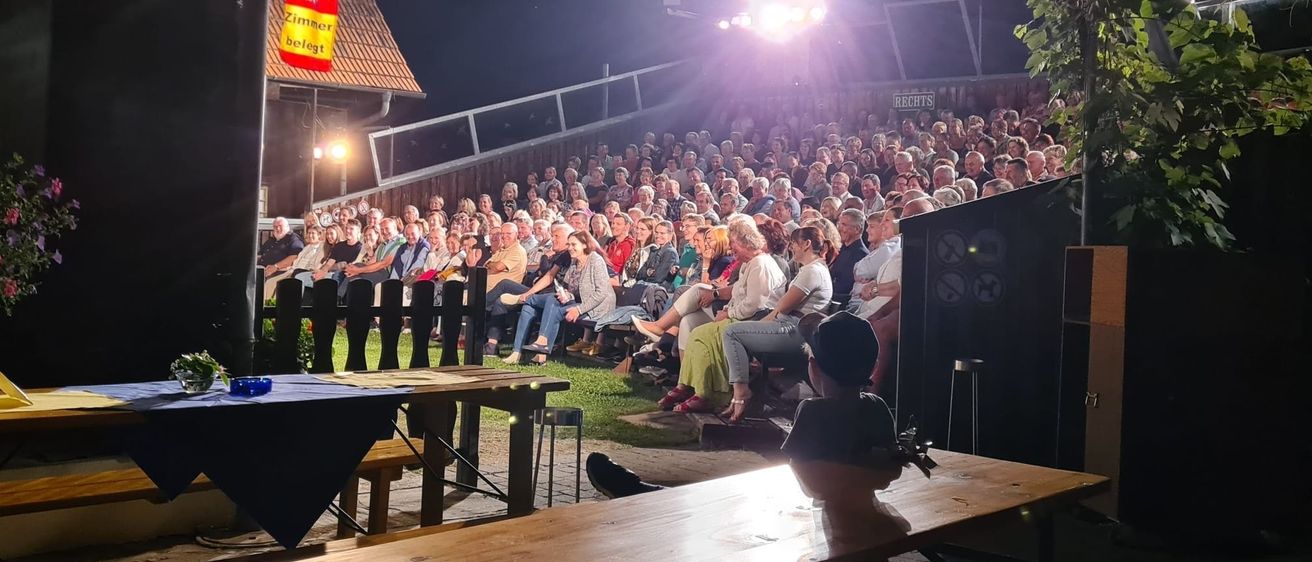 Eine große Menschenmenge sitzt bei einer nächtlichen Veranstaltung im Freien, mit einer Flagge und einer Bühne. Der Bereich ist gut beleuchtet von Scheinwerfern.