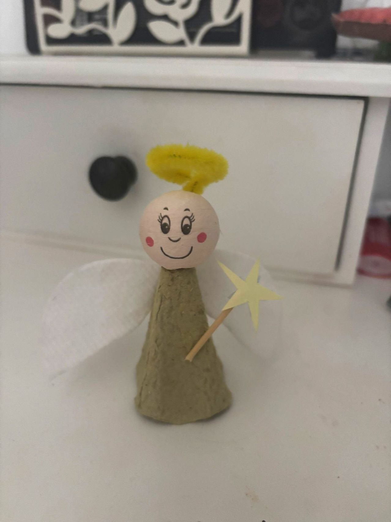 Eine kleine Holzfigur eines Engels mit gelbem Haar und Flügeln steht auf einer weißen Oberfläche. Sie hält einen gelben Sternenstab in der rechten Hand.