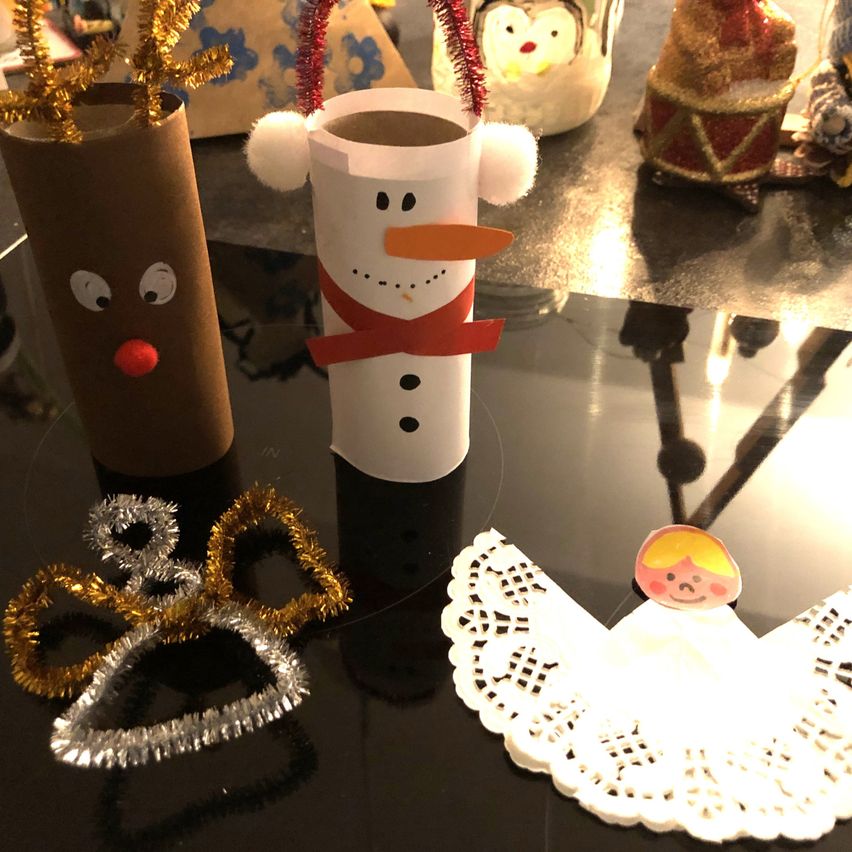Auf einem Tisch sind Weihnachtsbasteleien zu sehen: ein Schneemann mit rotem Band, ein brauner Rentier, ein Engel mit Flügeln und eine goldene Engelskopfbedeckung.