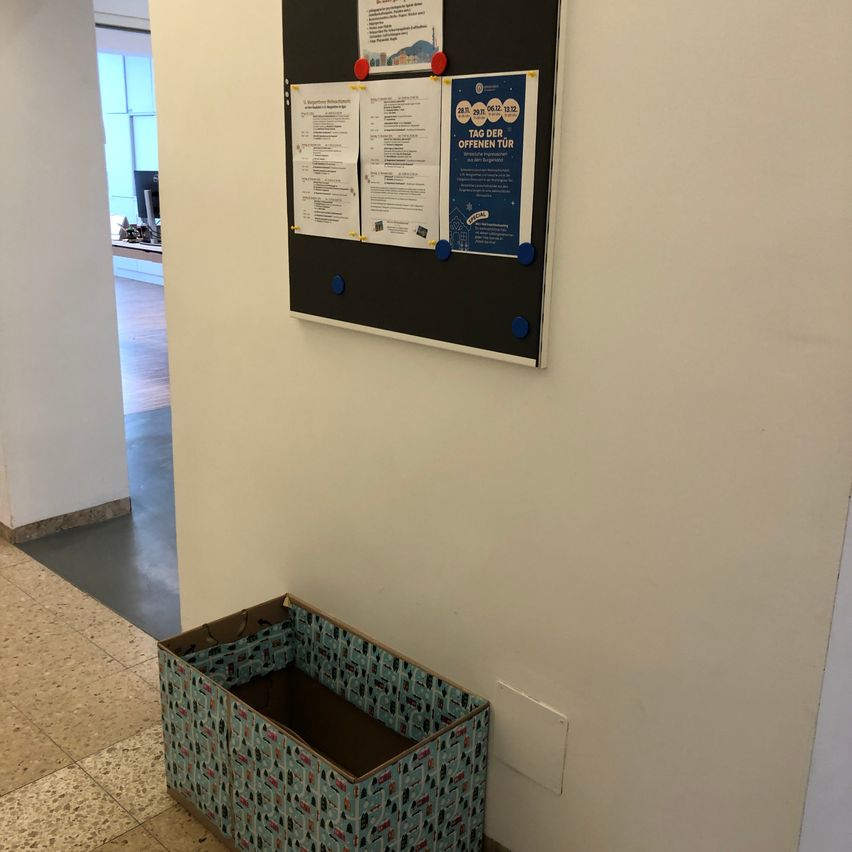 Ein Anschlagbrett mit verschiedenen angehefteten Papieren ist an einer Wand montiert. Darunter steht ein Aufbewahrungsbox auf dem Boden.