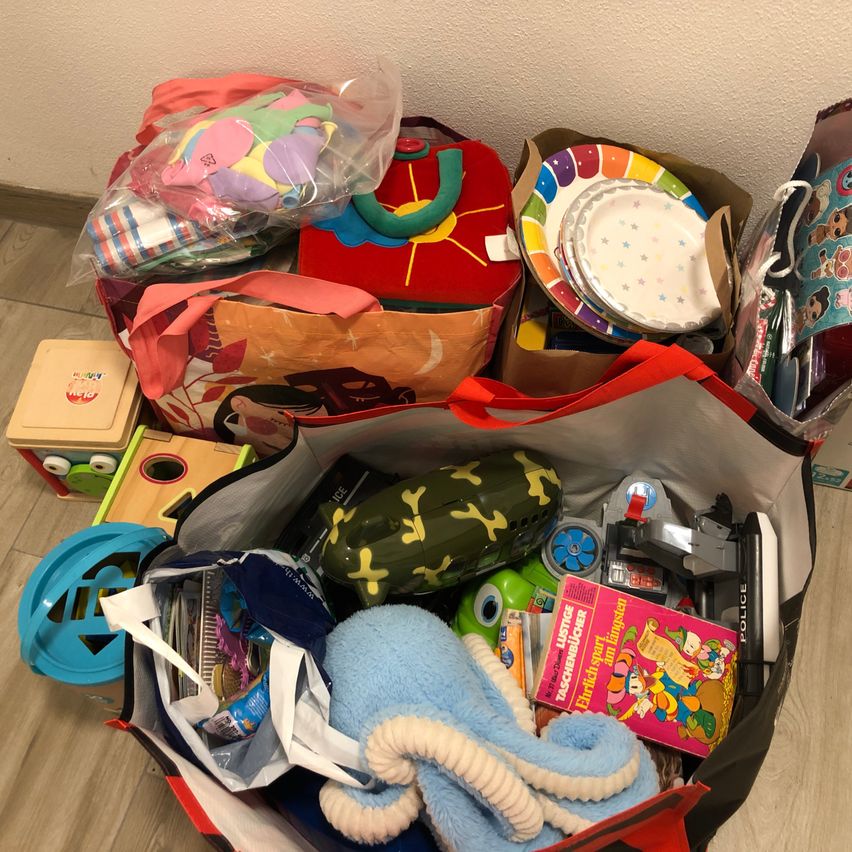 Verschiedene Gegenstände sind auf einem Holzboden ausgestellt, darunter Spielzeug, ein Buch und ein mit bunten Gegenständen gefüllter Beutel. Ein rote Kasten mit Sonnendesign ist ebenfalls sichtbar.