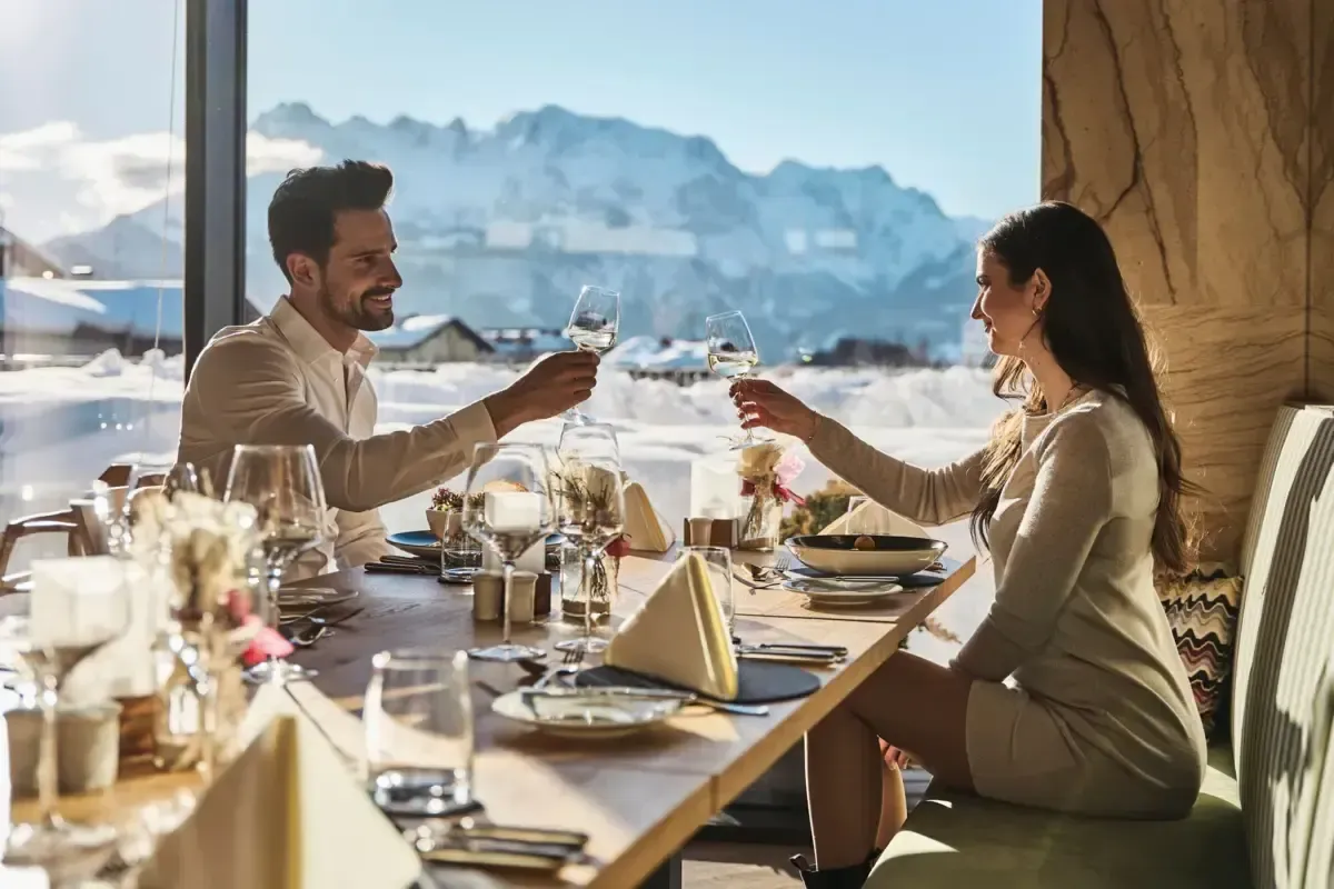 Ein Paar prostet mit Weingläsern in einem Restaurant mit Blick auf einen verschneiten Berg.