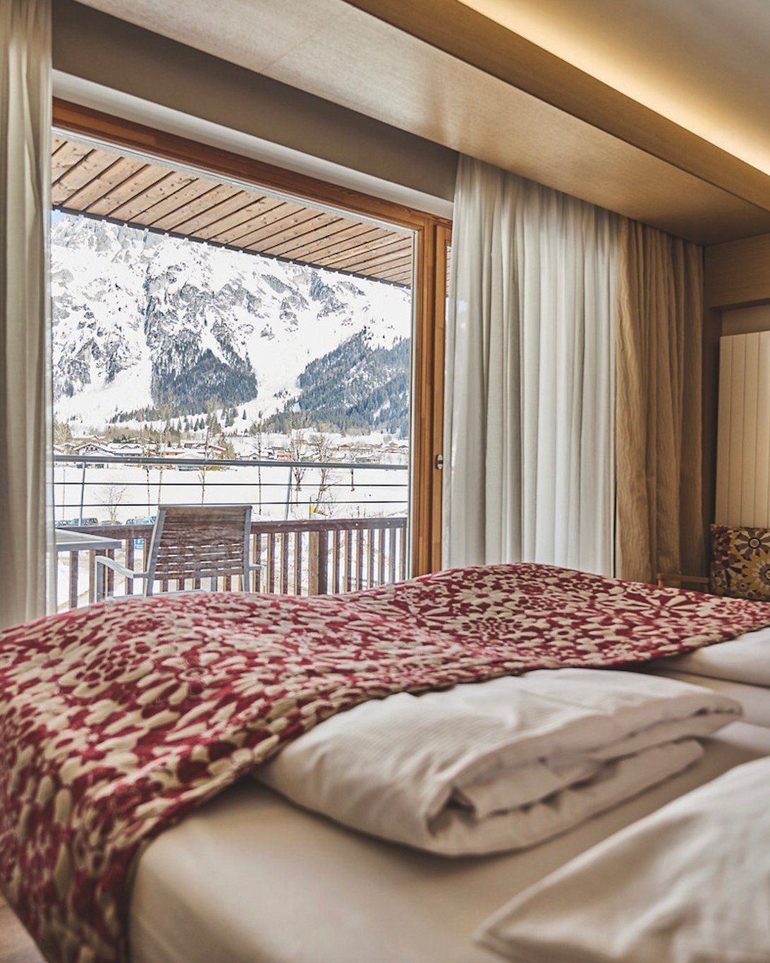 Ein ordentlich gemachtes Bett mit einer roten Blumendecke in einem Hotelzimmer. Das Zimmer hat Holzdecken und einen Balkon mit Blick auf verschneite Berge.