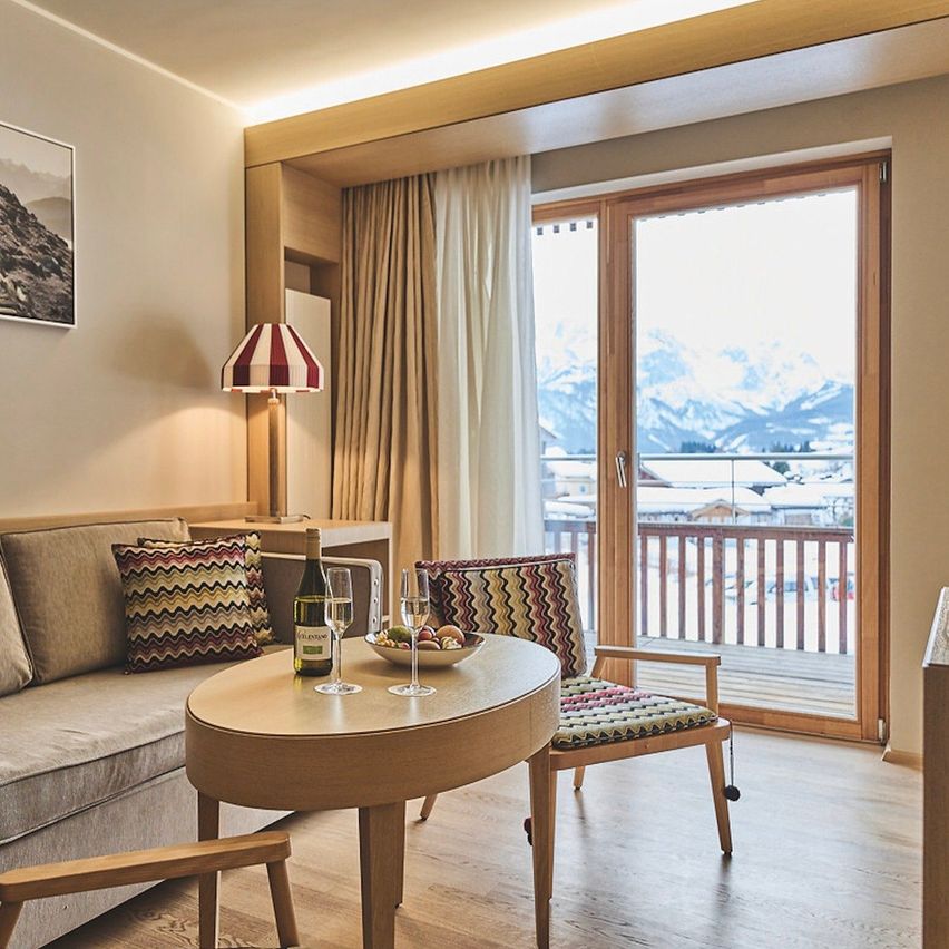 Ein gemütliches Wohnzimmer mit einem Sofa, einem Beistelltisch, einer Lampe und einem kleinen runden Tisch mit Weinläsern und einer Obstschale. Es gibt ein großes Fenster mit Blick auf einen Balkon und verschneite Berge.